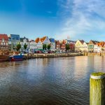 Blick auf das Flussufer mit farbenfrohen Gebäuden, Booten und Gehwegen unter einem strahlend blauen Himmel.