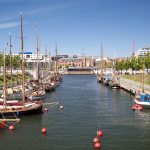 Segelboote liegen mit roten Bojen in einem Kanal vor Anker, umgeben von Bäumen und Gebäuden unter einem klaren blauen Himmel.