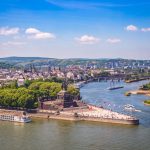 Luftaufnahme des Deutschen Ecks in Koblenz, Deutschland. Sie zeigt den atemberaubenden Zusammenfluss der Flüsse und die geschäftige Stadtlandschaft unter einem strahlend blauen Himmel, mit Booten, die anmutig durch die malerischen Gewässer navigieren.