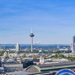 Luftaufnahme einer Stadtlandschaft mit einer Kathedrale, modernen Gebäuden und einem Fernsehturm unter einem klaren blauen Himmel.