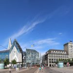 Panoramablick auf einen Stadtplatz mit modernen und historischen Gebäuden unter einem blauen Himmel.