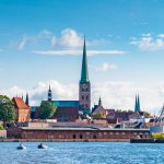 Panoramablick auf die Küstenstadt Lübeck mit Kirchtürmen, historischen Gebäuden, Booten und blauem Himmel.