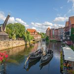 Historische Flusslandschaft in Lüneburg mit alten Gebäuden, Booten und leuchtenden Blumen. Sonniger Tag mit teilweise bewölktem Himmel.