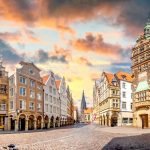 Der Stadtplatz von Münster strahlt mit seinen reich verzierten Gebäuden und dem majestätischen Glockenturm, der sich vor einem leuchtenden, bewölkten Abendhimmel abzeichnet, den Charme des historischen europäischen Stils aus.