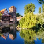 Malerische Aussicht auf ein historisches Fachwerkgebäude in Nürnberg an einem ruhigen Fluss, umgeben von üppigen grünen Bäumen und einem klaren blauen Himmel.