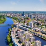 Eine Luftaufnahme von Offenbach zeigt seine reizvolle Stadtlandschaft mit dem glitzernden Fluss, der sich unter einer historischen Brücke hindurchschlängelt, umgeben von üppigem Grün unter einer strahlenden Sonne.