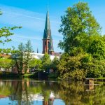 Eine heitere Parkszene in Oldenburg zeigt einen ruhigen Teich, üppiges Grün und einen Kirchturm, der sich im Hintergrund unter einem strahlend blauen Himmel erhebt.