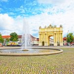 Ein historischer Bogen und Brunnen auf einem Platz mit Kopfsteinpflaster, umgeben von Gebäuden und Bäumen unter einem bewölkten Himmel.