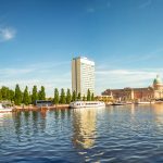 Panoramablick auf eine Stadtlandschaft am Flussufer in Potsdam mit Booten, Bäumen, einem Kuppelgebäude und einem modernen Turm.