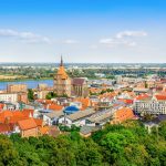 Panoramablick auf die Stadtlandschaft von Warnemünde mit roten Dächern, historischen Gebäuden und dem Fluss im Hintergrund unter klarem Himmel.