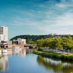 Die Stadtlandschaft von Saarbrücken erwacht mit einem Fluss zum Leben, der sich anmutig durch moderne Gebäude und üppige Bäume schlängelt, und das alles unter einem klaren, blauen Himmel.