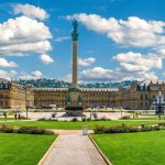 Ein großer Platz in Stuttgart mit einer zentralen Säule, umgeben von grünen Rasenflächen und historischen Gebäuden, lockt Reisende unter einem blauen Himmel mit Wolken.