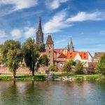 Panoramablick auf eine Stadt am Flussufer mit historischen Gebäuden, Bäumen und einem klaren blauen Himmel.