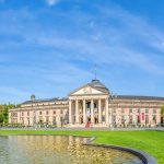 Panoramablick auf ein prächtiges Gebäude mit einem Brunnen, gepflegten Rasenflächen und einer Baumreihe unter einem blauen Himmel.