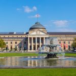 Das historische Wiesbadener Gebäude verfügt über einen großen Brunnen und einen gepflegten Rasen, elegant eingerahmt von Bäumen unter einem klaren blauen Himmel.