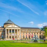 Ein großes historisches Gebäude mit Säulen und einer Kuppel steht majestätisch in Wiesbaden, umgeben von üppigen grünen Rasenflächen und einem eleganten Brunnen unter einem klaren blauen Himmel.