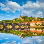 Malerische Aussicht auf eine historische Brücke in Würzburg mit farbenfrohen Gebäuden und einem Schloss auf einem Hügel, das sich im ruhigen Fluss spiegelt.