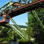 Eine orangefarbene Einschienenbahn überquert eine grüne Brücke über einen Fluss, umgeben von üppigen Bäumen.