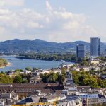 Panoramablick auf Bonn mit dem Rhein, historischen Gebäuden und Hügeln in der Ferne unter einem teilweise bewölkten Himmel.