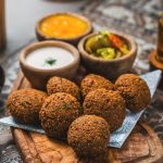 Falafelbällchen auf einer Holzplatte mit Schüsseln voller Soße, eingelegtem Gemüse und Gemüse, auf einer gemusterten Tischdecke angerichtet.