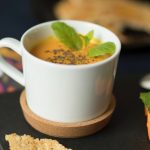 Eine weiße Tasse Kürbissuppe, garniert mit Minzblättern und -samen auf einem Korkuntersetzer, mit Brot daneben.