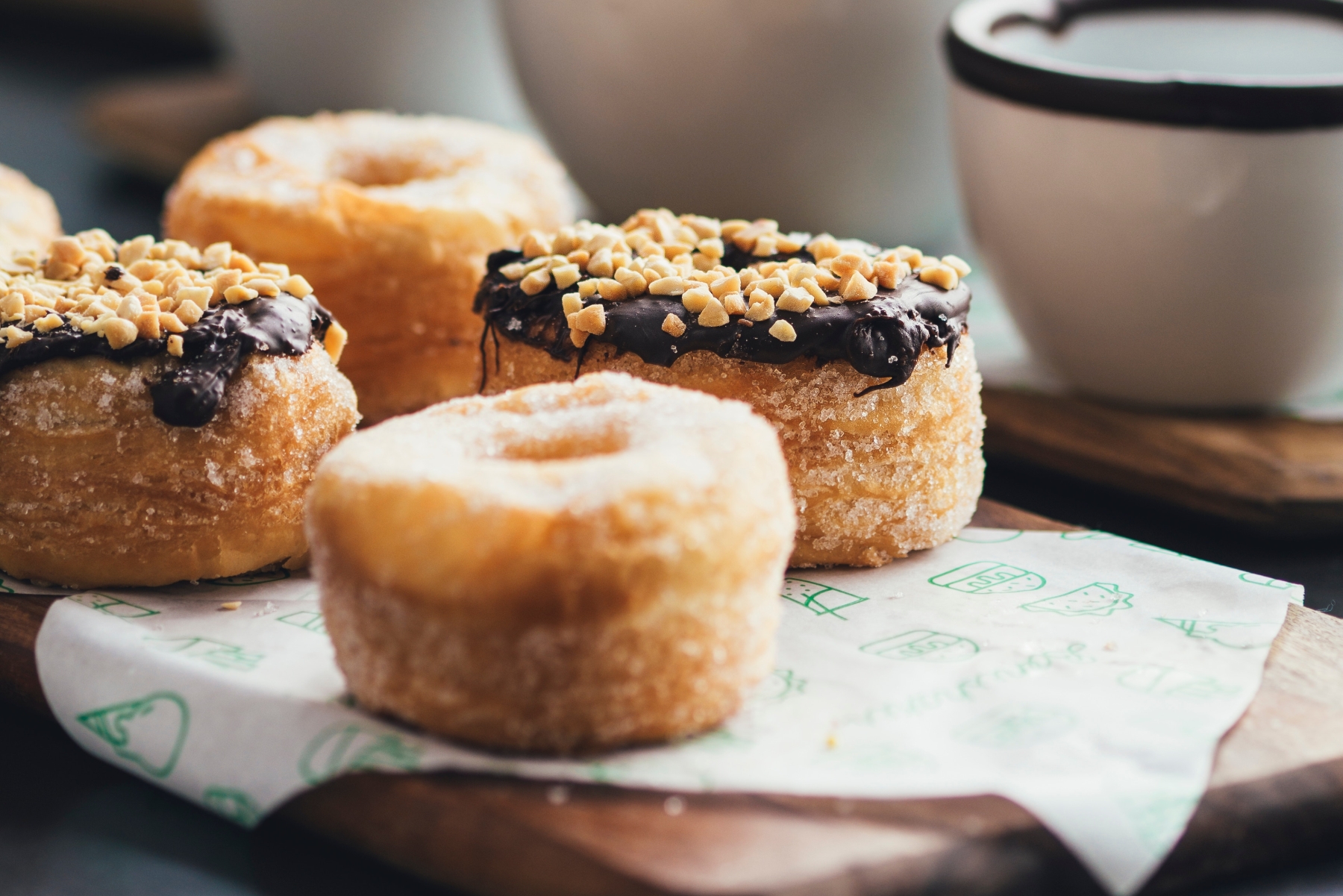 Nahaufnahme von mit Schokolade und Nüssen garnierten Cronuts auf einem Holztablett, die die gemütliche Atmosphäre einer Weihnachtsfeier heraufbeschwören, mit einer Tasse im Hintergrund.