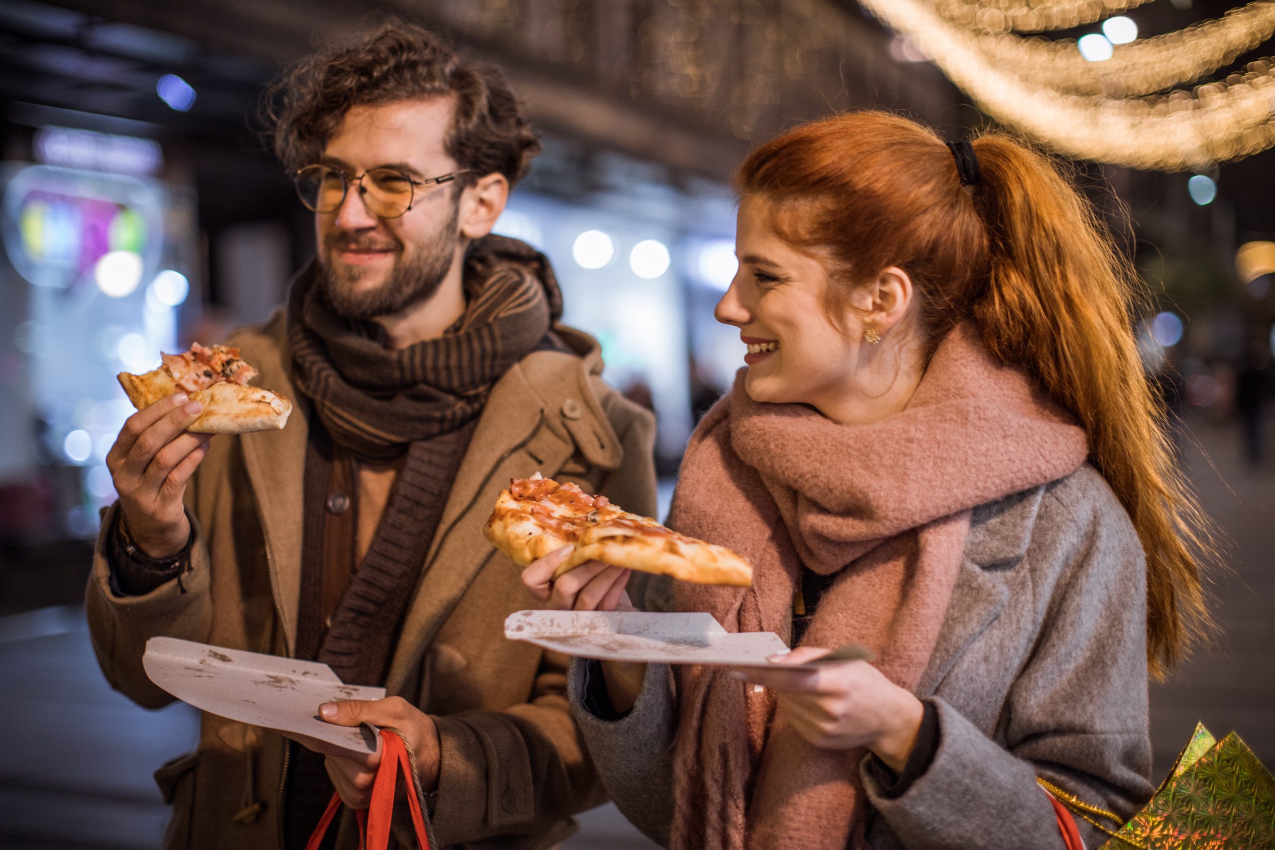 Zwei Menschen lächeln und genießen Pizza, während sie eine festlich beleuchtete Straße entlangschlendern und die fröhliche Stimmung einer Weihnachtsfeier einfangen.