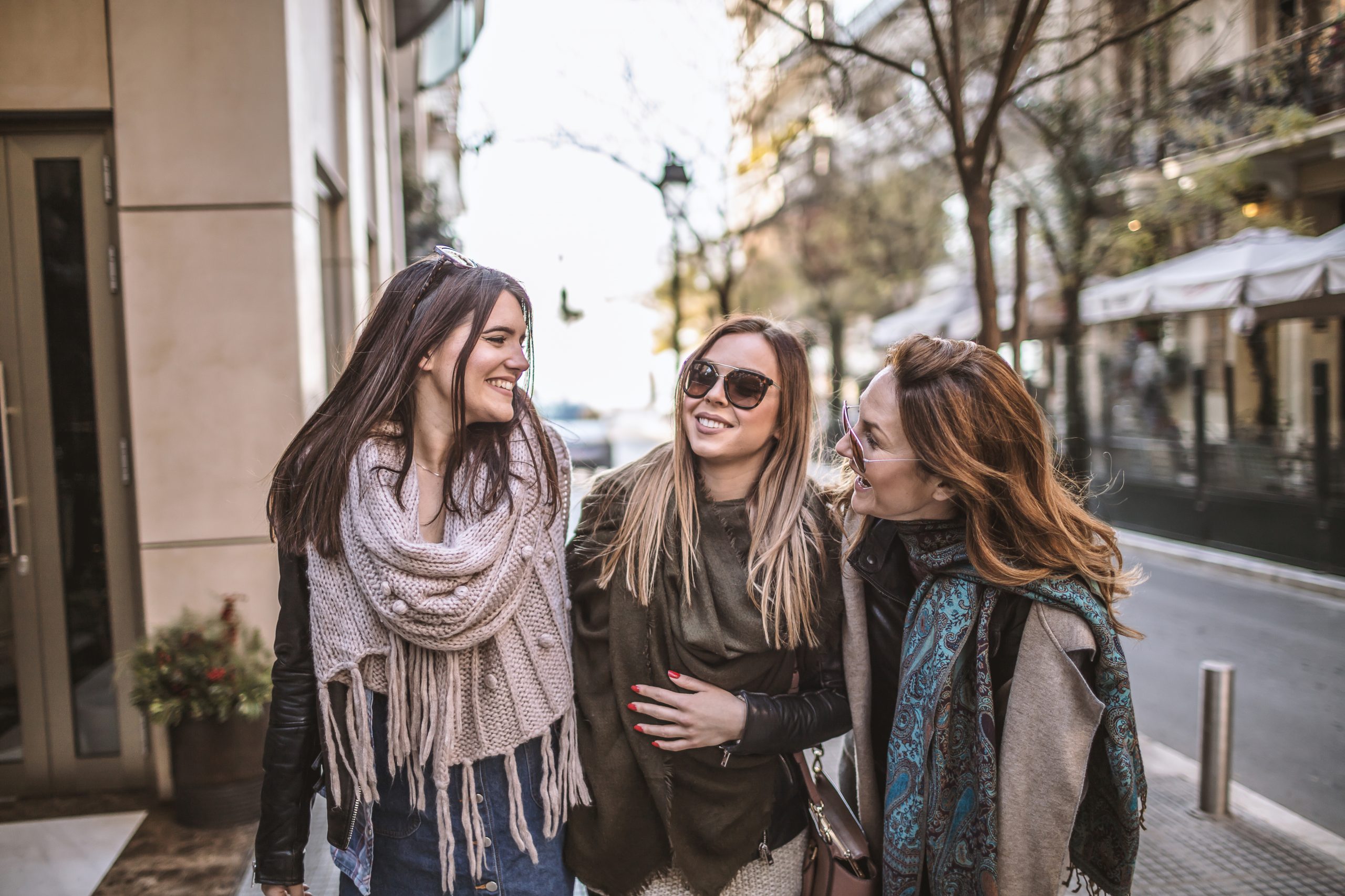 Drei Frauen, die lächelnd Arm in Arm mit Schals und Sonnenbrillen durch eine Stadtstraße gehen, genießen ihren Tag im festlichen Geist einer Weihnachtsfeier.