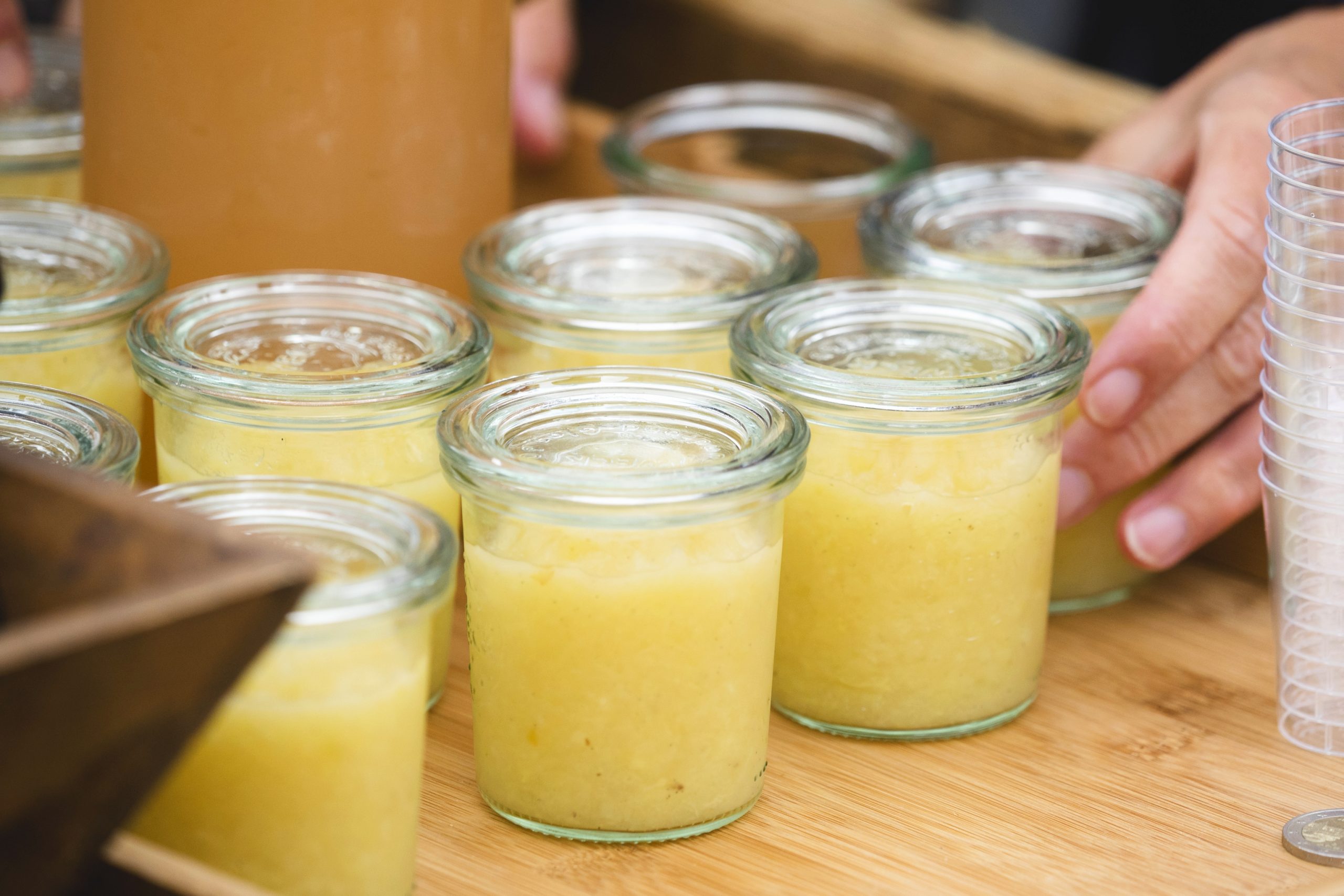 Gläser mit gelbem Smoothie stehen auf einem Holztisch und sind bereit, bei der Weihnachtsfeier genossen zu werden. Hände greifen nach den durchsichtigen Plastikbechern, die daneben stehen.