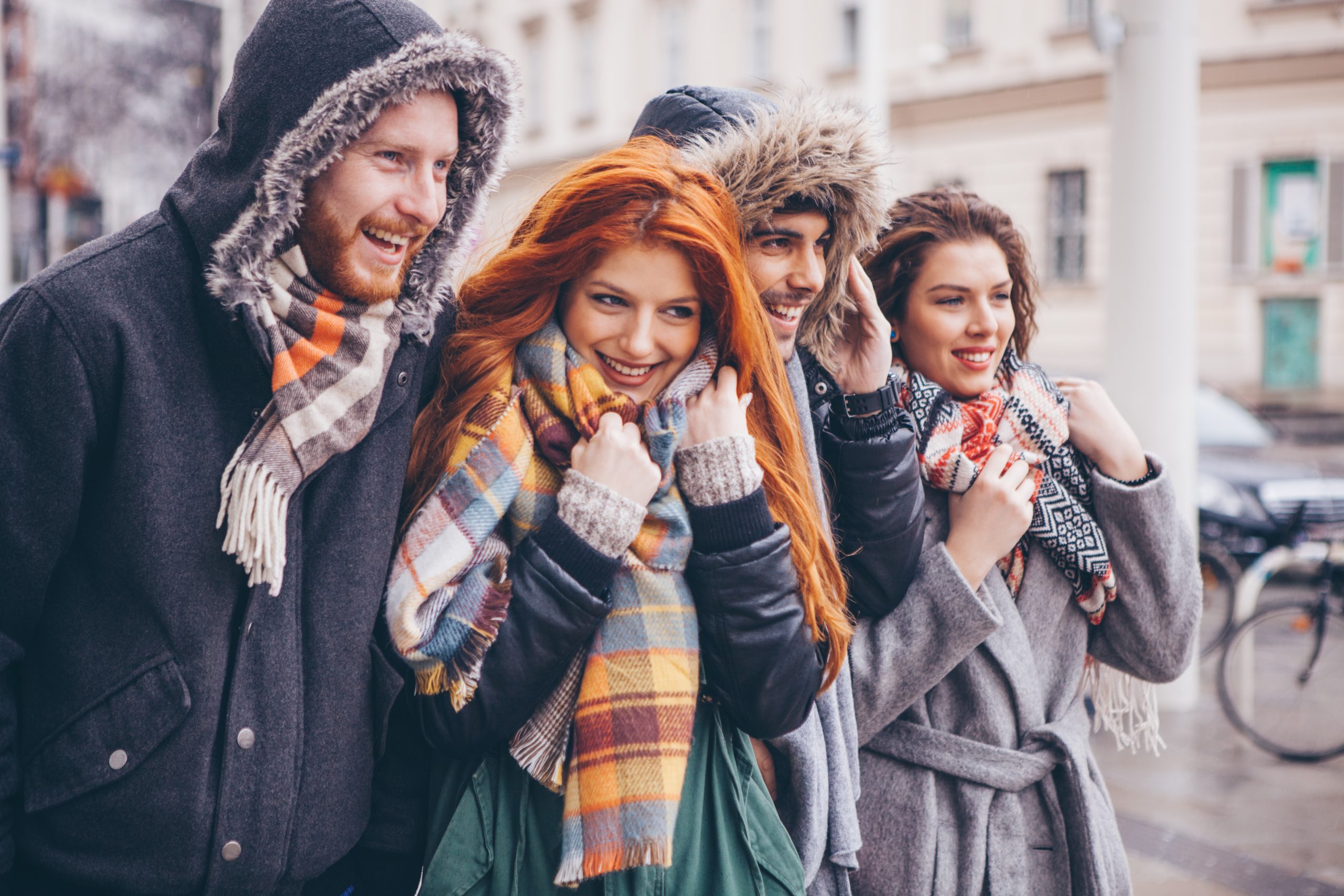 Vier Freunde in warmen Mänteln und Schals, die an einem kühlen Tag lächelnd im Freien spazieren gehen, vielleicht auf dem Weg zu einer festlichen Weihnachtsfeier.