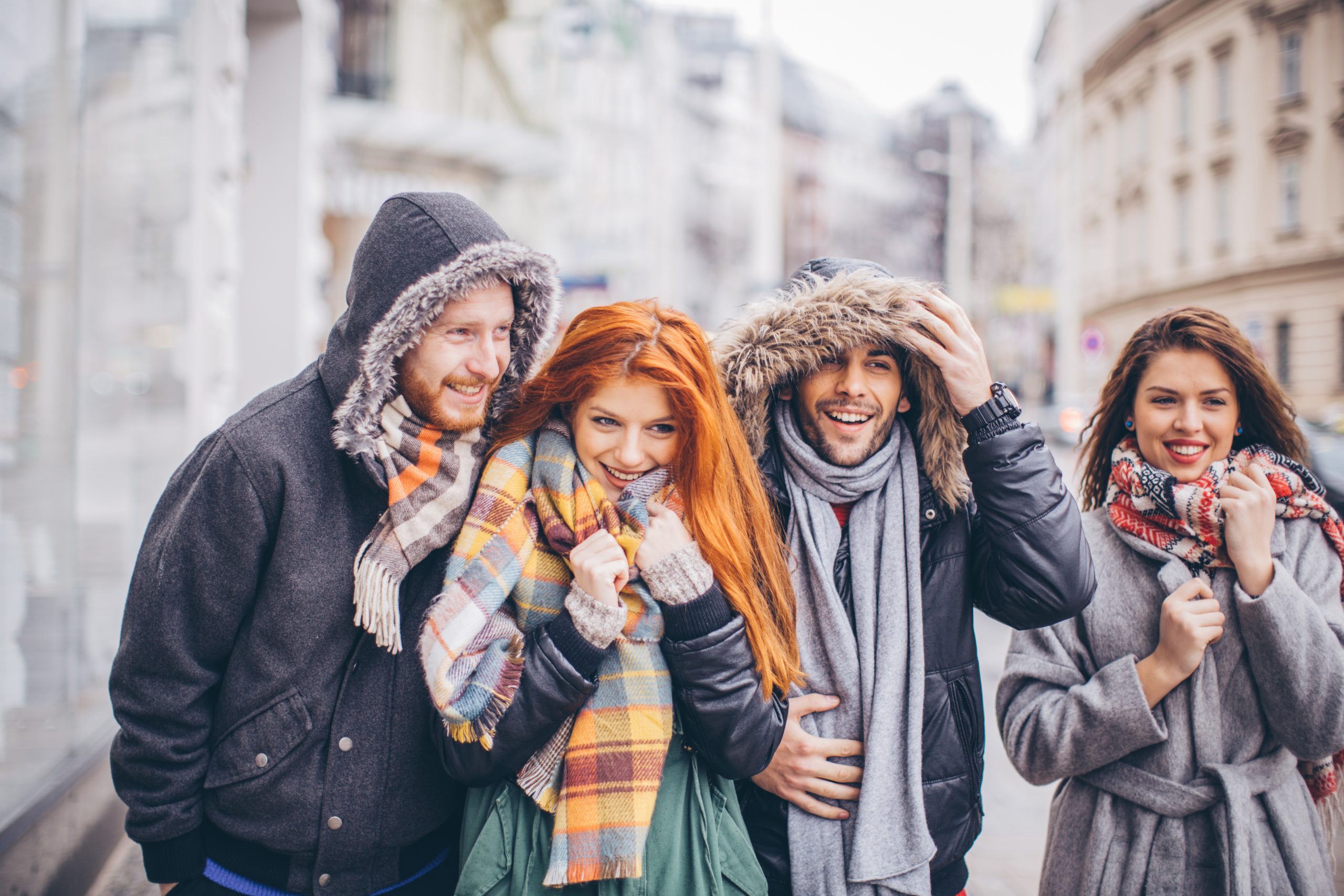 Vier in warme Mäntel und Schals eingemummelte Menschen lächeln, während sie an einem kühlen Tag durch eine Stadtstraße schlendern. Ihr Lachen lässt die Vorfreude auf die bevorstehende Weihnachtsfeier erahnen.