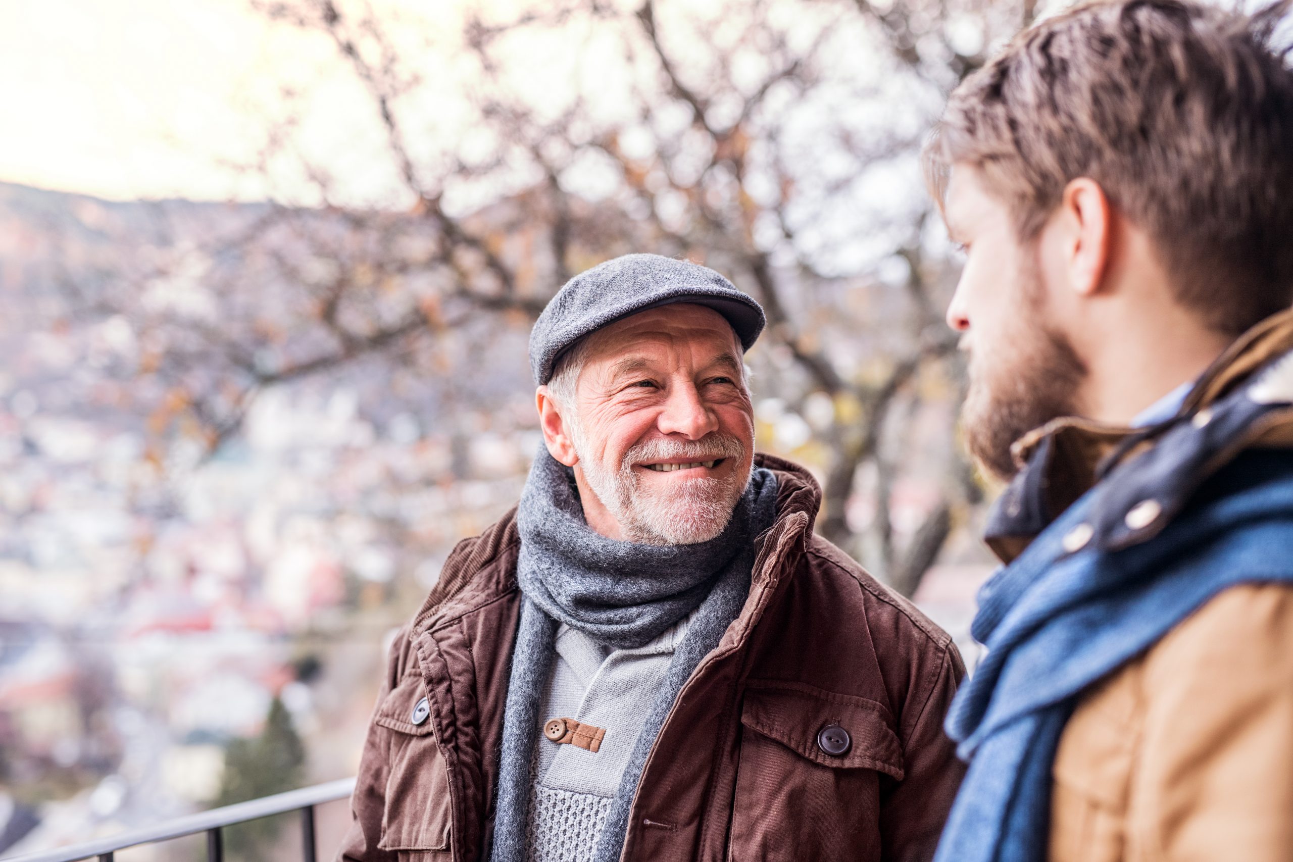 Zwei Männer in warmer Kleidung unterhalten sich draußen bei einer Weihnachtsfeier. Einer lächelt und trägt eine Schiebermütze. Unscharfe Bäume unterstreichen die festliche Kulisse.