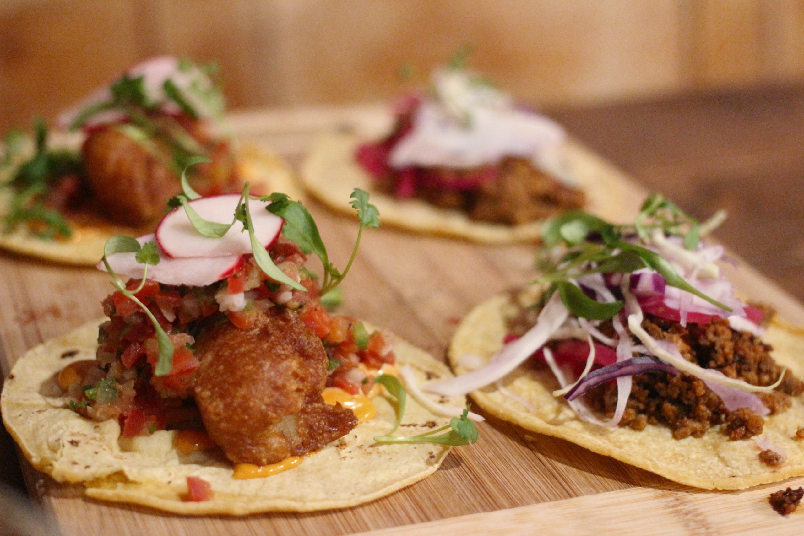 Vier verschiedene Tacos mit unterschiedlichen Belägen, darunter Radieschenscheiben und -grün, sind hübsch auf einer Holzoberfläche angerichtet und versprühen den festlichen Charme einer Weihnachtsfeier.