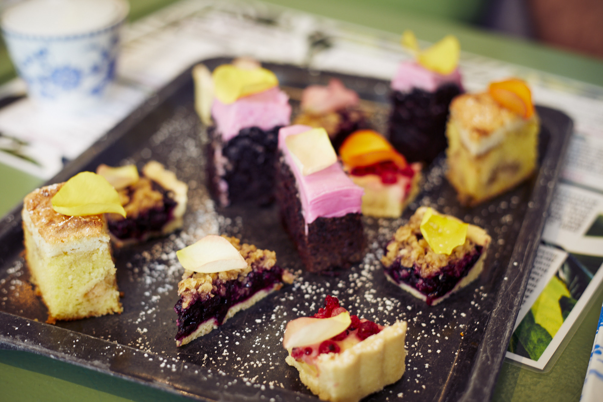 Ein Tablett mit verschiedenen bunten Backwaren, Kuchen und Törtchen, garniert mit essbaren Blütenblättern, steht elegant auf einem grünen Tisch, perfekt für eine festliche Weihnachtsfeier.