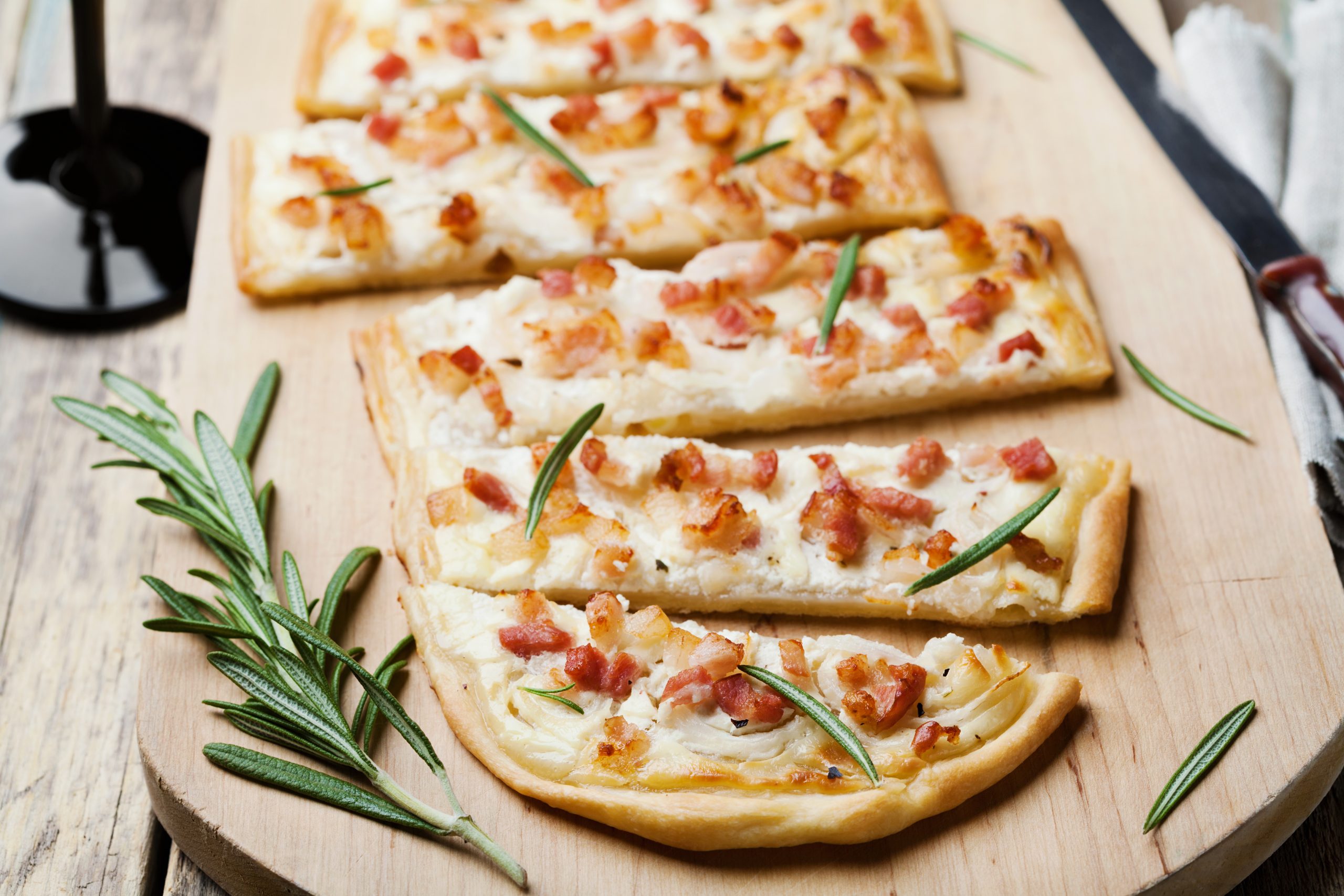 Mit Speck, Käse und Rosmarin belegtes Fladenbrot liegt verführerisch auf einem Holzbrett, perfekt gepaart mit einem Glas Rotwein – ideal zum Genießen bei Ihrer Weihnachtsfeier.