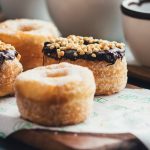 Mit Schokolade und Nüssen garnierte Cronuts auf einem Holzbrett neben einer Tasse Kaffee sind der perfekte festliche Leckerbissen für Ihre Weihnachtsfeier.