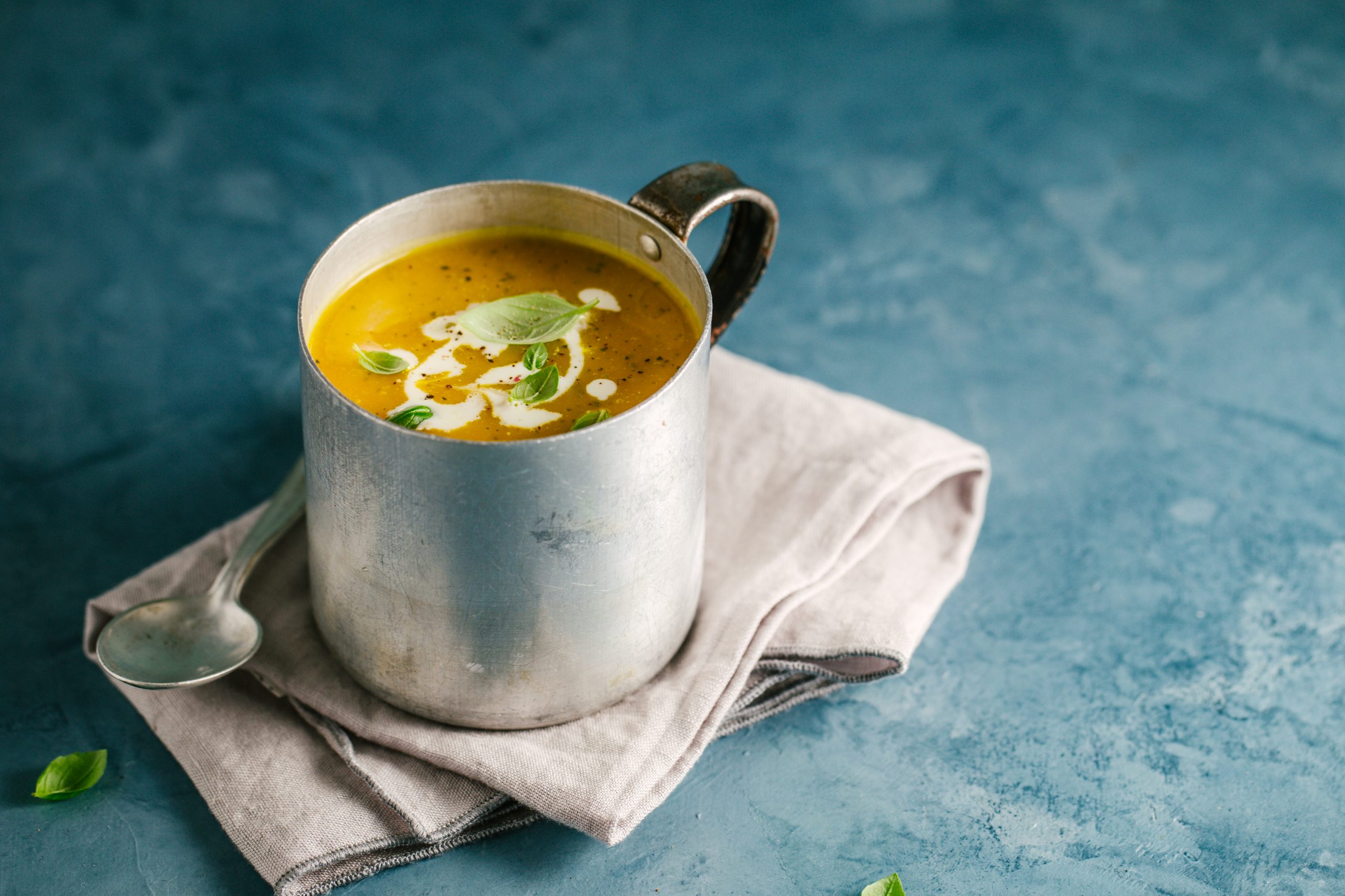 Eine rustikale Tasse cremiger Kürbissuppe mit Kräutern steht auf einer gefalteten Serviette und erinnert an die Wärme einer Weihnachtsfeier. Dazu kommt ein Löffel auf einer blauen Oberfläche.