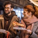 Ein lächelndes Paar genießt beim Spaziergang im Freien, in Schals und Mäntel gehüllt, Pizza, im Hintergrund funkeln die festlichen Lichter einer Weihnachtsfeier.