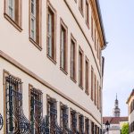 Historische Gebäude mit reich verzierten Fenstern säumen eine Straße. Im Hintergrund ist ein Turm mit einer Kuppel zu sehen.