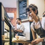 Eine Frau in einer Schürze serviert Essen aus einer Vitrine in einem Café und lächelt, im Hintergrund ist ein männlicher Barista zu sehen.