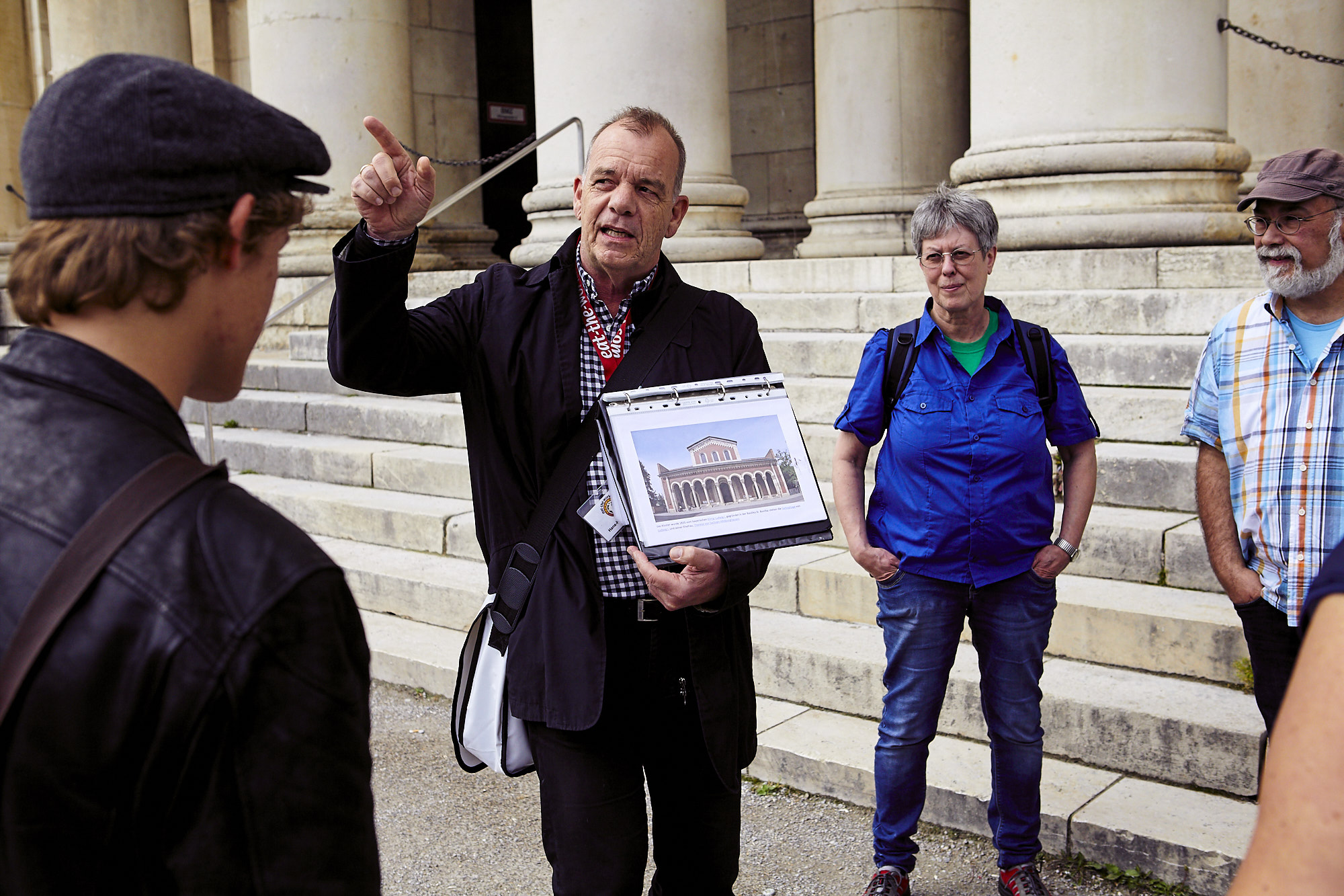 Ein Reiseführer hält das Bild eines Gebäudes in der Hand und spricht zu einer Gruppe vor Steinsäulen und -stufen, die an die historische Architektur Augsburgs erinnern.