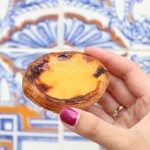 Eine Hand, die ein Pastel de Nata hält, mit den leuchtend blau und orange gemusterten Kacheln im Hintergrund, ergibt ein Bild, das genauso reizvoll ist wie das Einlösen eines Gutscheins für Ihre Lieblingssüßigkeit.