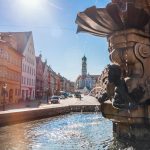 Brunnen mit Engelsstatue auf einem sonnigen Stadtplatz; historische farbenfrohe Gebäude säumen die Straße.