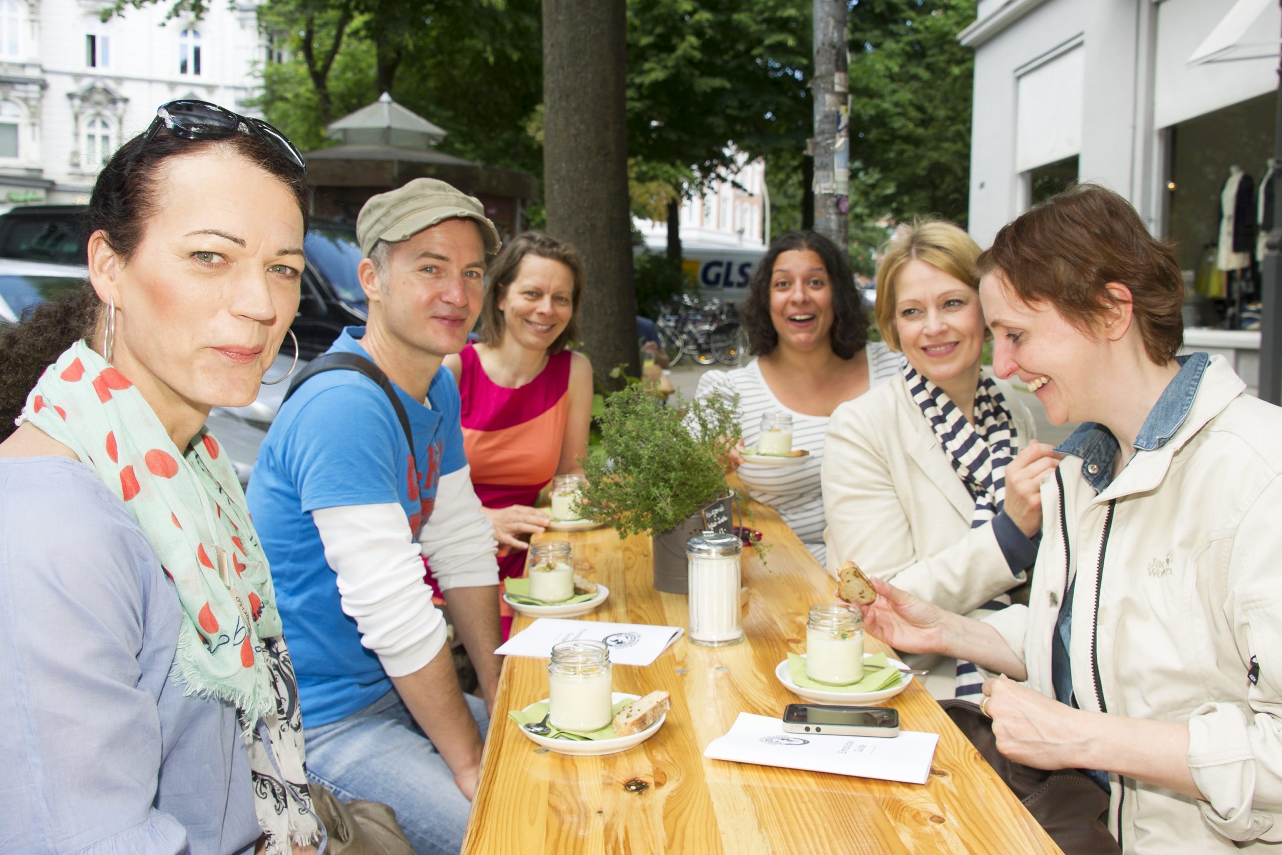 Eine Gruppe von Kollegen sitzt an einem Café-Tisch im Freien, lächelt und genießt während ihres Betriebsausflugs Getränke.