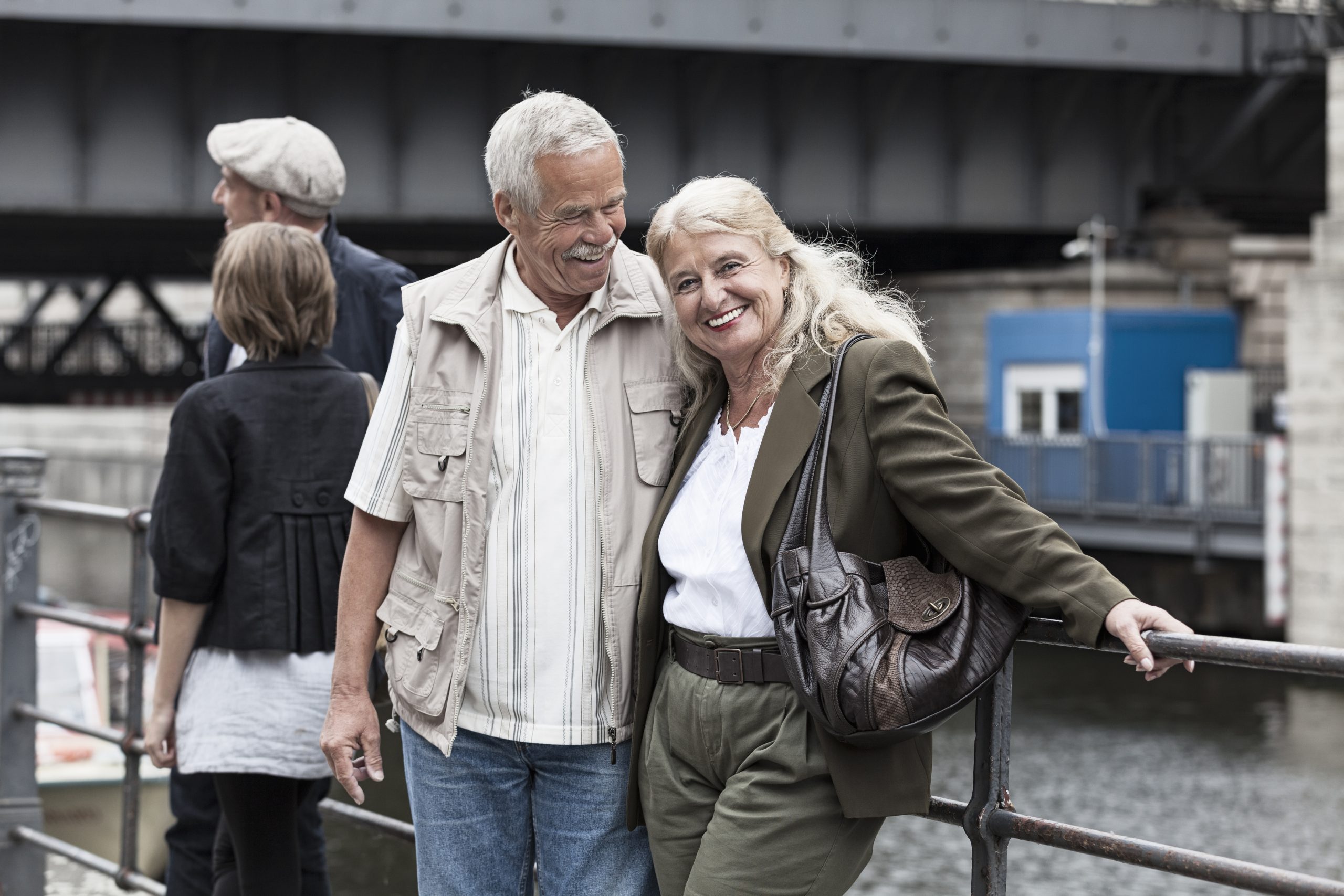 Lächelndes Paar steht an einem Ufergeländer, im Hintergrund eine Brücke – eine ideale Kulisse, um Erinnerungen festzuhalten oder eine einzigartige Geschenkidee zu finden. Andere Menschen tragen zur lebendigen Szene bei.