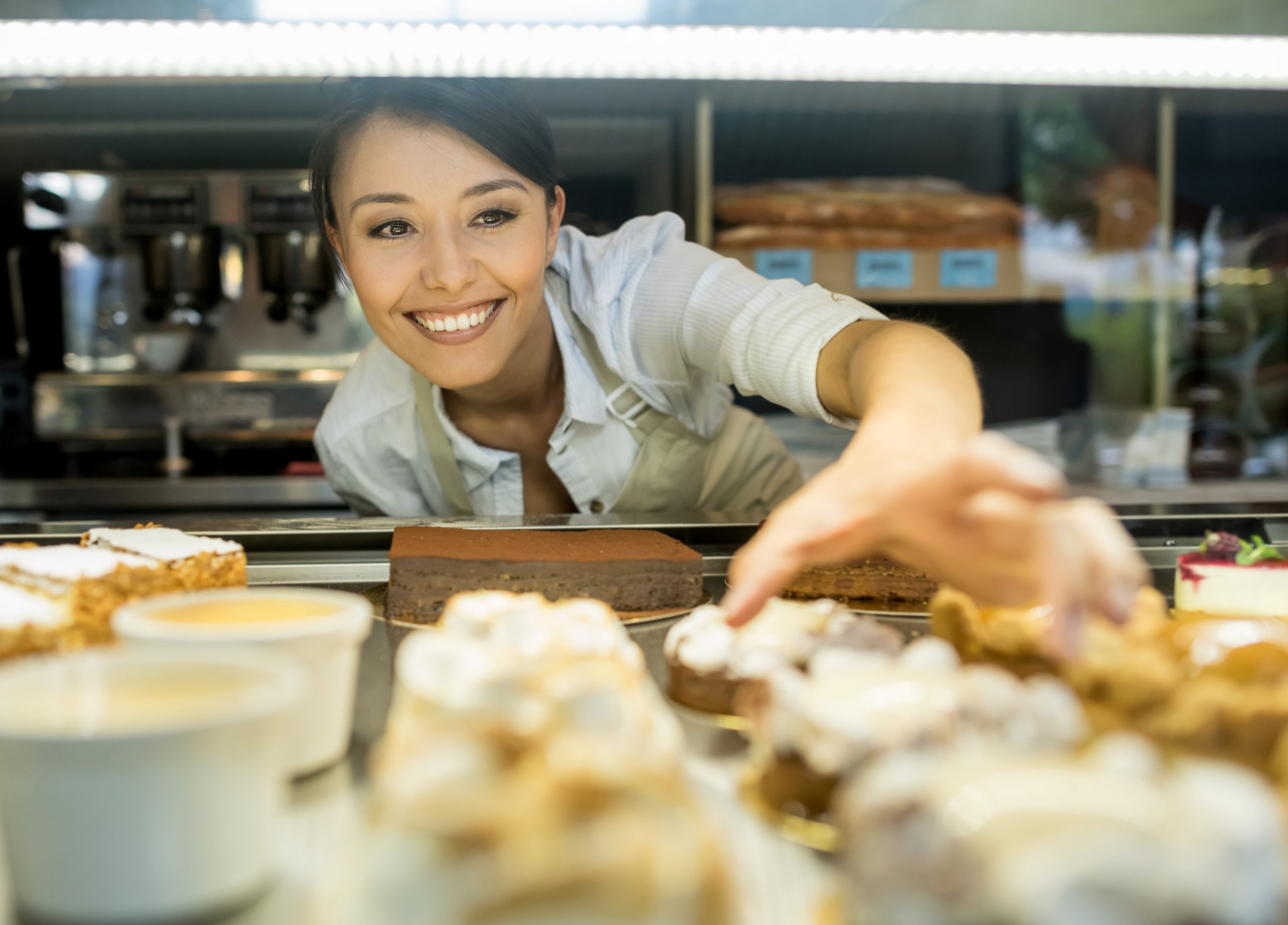 Eine Frau steht fröhlich hinter der Theke einer Bäckerei und arrangiert fachmännisch Gebäck und Kuchen – jedes davon eine entzückende Geschenkidee, die garantiert Freude bereitet.