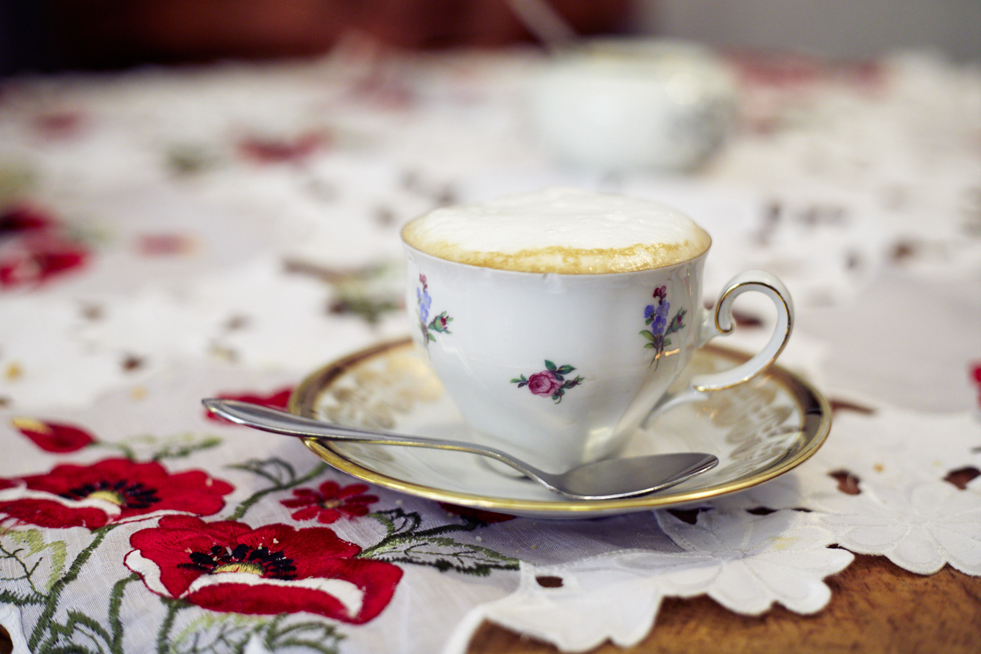 Eine geblümte Porzellantasse mit schaumigem Cappuccino ruht auf einer Untertasse und einem Löffel auf einer bestickten Tischdecke und schafft die perfekte Kulisse für eine gemütliche Weihnachtsfeier.