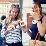 Zwei Frauen essen im Freien ein Eis, lächeln und stehen auf einem gemusterten Bürgersteig mit Gebäuden im Hintergrund.