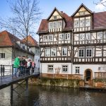 Menschen stehen an einem sonnigen Tag auf einer Brücke vor einem historischen Fachwerkhaus an einem Fluss.