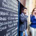 Eine Kreidetafel mit Menü im Freien, im Hintergrund zwei Personen, die aus Gläsern trinken.